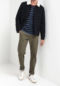 Herrjacka i svart med vit pälskrage, marinblå randig skjorta, olivgröna byxor och vita sneakers. Enkel design med knappstängning.