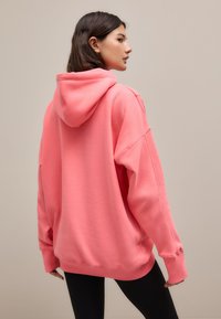 Roza jopa s kapuco, sproščenega kroja z rebrastimi manšetami in spodnjim robom. Mehka tekstura, s široko kapuco in spuščenimi rameni.