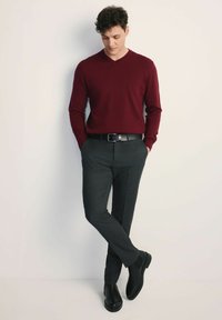 Bordowy sweter w serek zestawiony z ciemnoszarymi spodniami o dopasowanym kroju i czarnymi skórzanymi botkami za kostkę, stojący na neutralnym tle.