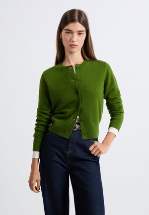 Cardigan in lana verde con scollatura tonda e chiusura con bottoni. Maniche lunghe e polsini a coste. Indossato sopra una maglietta bianca e abbinato a jeans scuri.