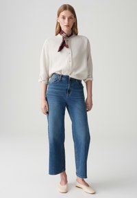 Camicia bianca a maniche lunghe, jeans blu a vita alta e ballerine color crema, con una sciarpa bordeaux intorno al collo per un tocco di dettaglio in più.