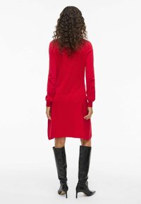 Rotes Langarmkleid mit gerader Silhouette, aus Strickstoff gefertigt, mit seitlichen Schlitzen, kombiniert mit knielangen schwarzen High Heels.