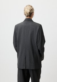Blazer à rayures gris foncé avec une coupe structurée, fente unique à l'arrière et poignets à trois boutons. Le tissu a une texture lisse.