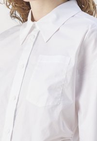 Gros plan du buste supérieur portant une chemise blanche à boutons avec col pointu et poche poitrine, cheveux bouclés châtain clair visibles.