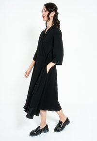 Fekete, wrap-stílusú ruha háromnegyedes ujjakkal, övvel a derékon és oldalsó zsebekkel. Fényes fekete slip-on cipővel van párosítva.