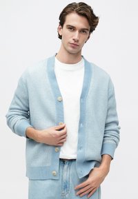 Mlad moški nosi svetlo modro zapnjen cardigan preko bele srajce in svetlo modrih kavbojk, medtem ko z eno roko prilagaja cardigan.