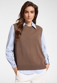 Maglione marrone senza maniche con orlo a costine, indossato sopra una camicia azzurro chiaro con bottoni ai polsini. Texture liscia, design casual.