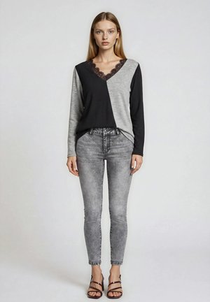 Kobieta nosząca czarną i szarą bluzkę z długim rękawem i dekoltem w szpic z koronkowym detalem, szare wyblakłe jeansy i brązowe sandały na paski, stojąca na jednolitym tle.