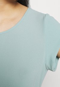 Ljus teal kortärmad topp gjord av texturerad tyg, med rundad halsringning och subtil sömnad längs kanterna.