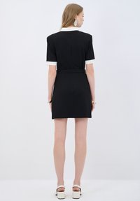 Schwarzes Kleid mit kurzen Ärmeln, weißen Bündchen, tailliertem Schnitt und geradem Saum. Glattes Material mit einem Gürteldetail an der Taille.