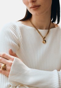 Goldene Halskette mit Herzanhänger, kombiniert mit einem goldenen Ring, getragen über einem weißen, gerippten Pullover, der eine glatte Textur und subtilen Glanz zeigt.