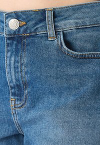 Jeans in denim di un blu medio con cuciture arancioni. Caratterizzati da una chiusura con bottone in metallo e una tasca frontale. Texture liscia.