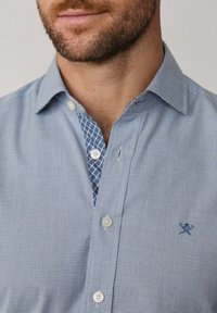 Muž v modro-bielej kockovanej košeli na gombíky s kontrastným vnútorným golierom a malou vyšitou nášivkou na hrudi.