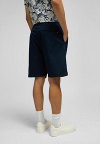 Marineblaue Shorts aus glattem Material mit seitlichen Taschen, kombiniert mit weißen Sneakern und gerippten Socken, von hinten gezeigt.