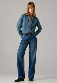 Ženska v dolgi kavbojkasti srajci z dolgimi rokavi, širokih kavbojkah z visokim pasom, črnem pasu in črnih gležnjarjih z visokimi petami stoji z rokami za hrbtom.