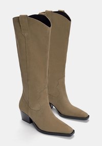 Beżowe zamszowe kowbojki w stylu western, z wąskim noskiem, blokowym obcasem i dekoracyjnym przeszyciem wzdłuż cholewki; wysoka budowa z bocznymi panelami.