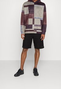 Mikina s kapucí v patchworkovém designu, obsahující různé odstíny fialové a šedé. Kombinováno s černými šortkami a černými teniskami.