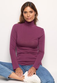 Frau mit braunen Haaren, die einen lila Rollkragenpullover, blaue Jeans und weiße Sneakers trägt, sitzt im Schneidersitz gegen eine weiße Wand.