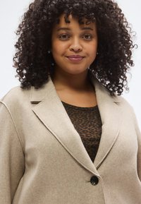 Παλτό μπεζ από μείγμα μαλλιού με γιακά με εγκοπές, ένα μαύρο κουμπί και απαλή υφή, φορεμένο πάνω από καφέ τοπ με λεοπάρ σχέδιο.