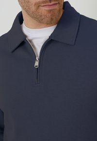 Marineblauer Quarter-Zip-Pullover aus glattem Stoff mit einem grauen Reißverschluss, ausgestattet mit einem umlegbaren Kragen und einer weißen Schicht darunter.