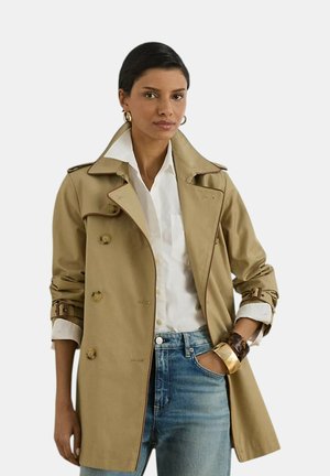 Donna che indossa un trench beige sopra una camicia bianca e jeans blu, accessoriata con orecchini a cerchio dorati e braccialetti spessi.