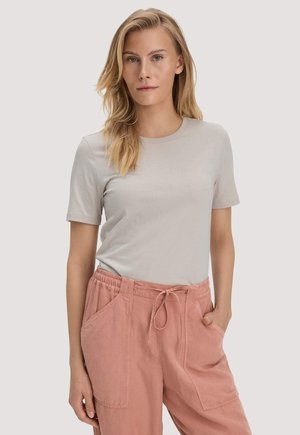 Blonde vrouw met los haar, gekleed in een lichtgrijs shirt met korte mouwen en roze broek met trekkoord, staand met één hand in de zak.