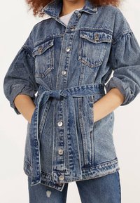 Jeansjacka med bälte i midjan, två bröstfickor, silverfärgade tryckknappar, ljusblå tvätt och lätt puffade ärmar.