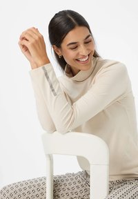 Lächelnde Frau mit dunklen Haaren in einem beigen Langarmoberteil mit Strassdetails an den Ärmeln, auf einem weißen Stuhl sitzend, mit gemusterten Hosen.