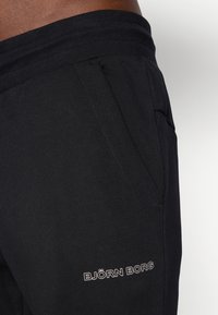Close-up of black sweatpants with elastic waistband and side pocket, featuring white "BJÖRN BORG" logo on thigh.