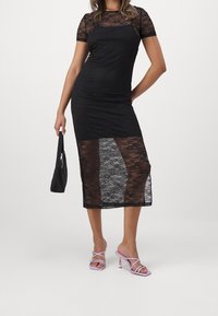 Kvinna iklädd en svart spetsklänning med genomskinliga kjoldelar, håller en liten svart handväska och bär lavendelfärgade sandaletter med hög klack.