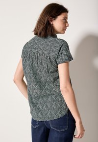 Camicetta a maniche corte di colore verde scuro con un intricato motivo a foglie bianche. Presenta un retro plissettato e un orlo arrotondato. Indossata con jeans scuri.
