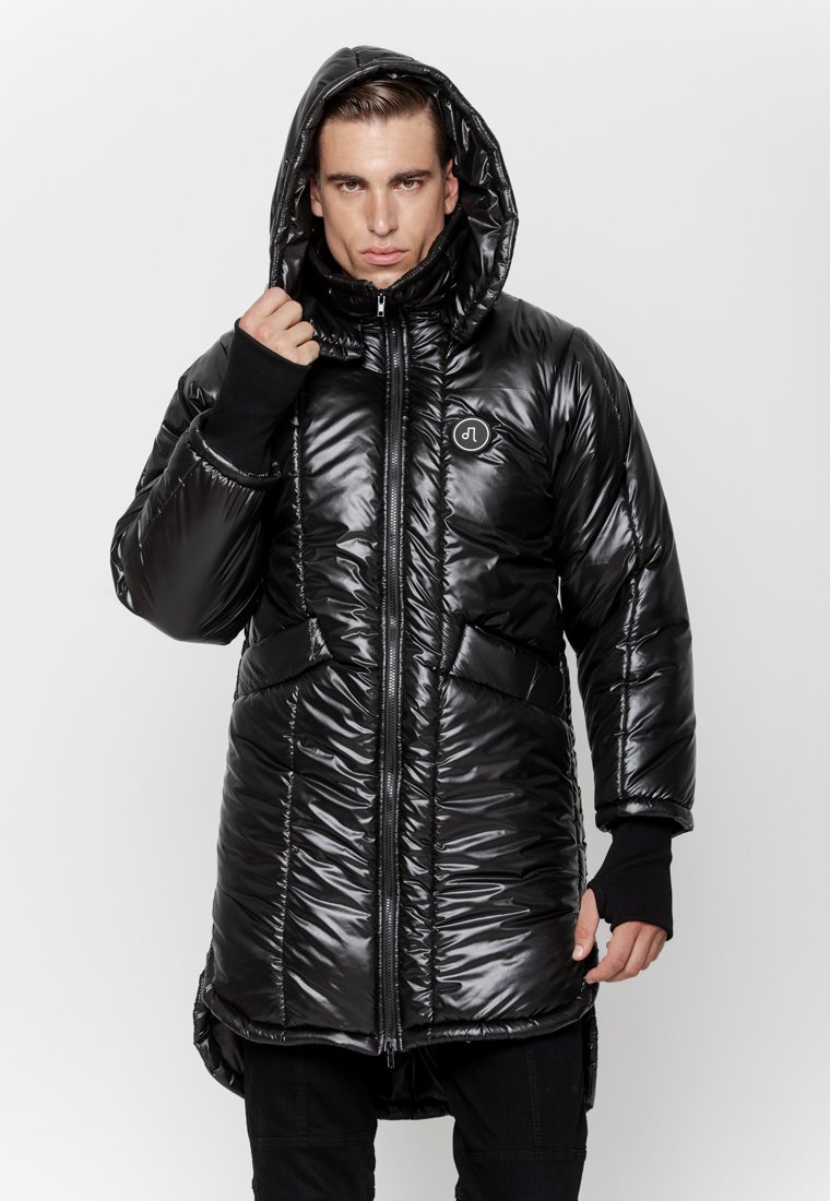 Mężczyzna w długiej, czarnej, błyszczącej kurtce puchowej z kapturem, trzymający kaptur jedną ręką, stojący na tle jasnego, jednolitego tła.