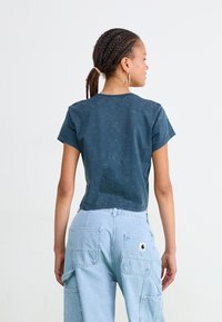 Short-sleeve dark teal t-shirt with a relaxed fit and faded texture, paired with light blue cargo pants featuring back pockets and logo patch.