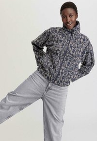 Fleece jack met hoge kraag, voorzien van een blauw en grijs abstract patroon, ritssluiting en elastische zoom, gecombineerd met lichtgrijze jeans.