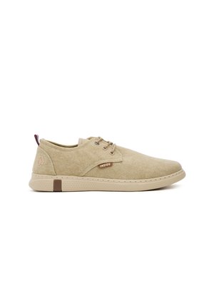 Zapatillas de lona beige con una textura suave, forma redondeada, diseño de cordones y acentos marrones, que presentan un logotipo y una cinta en el talón.