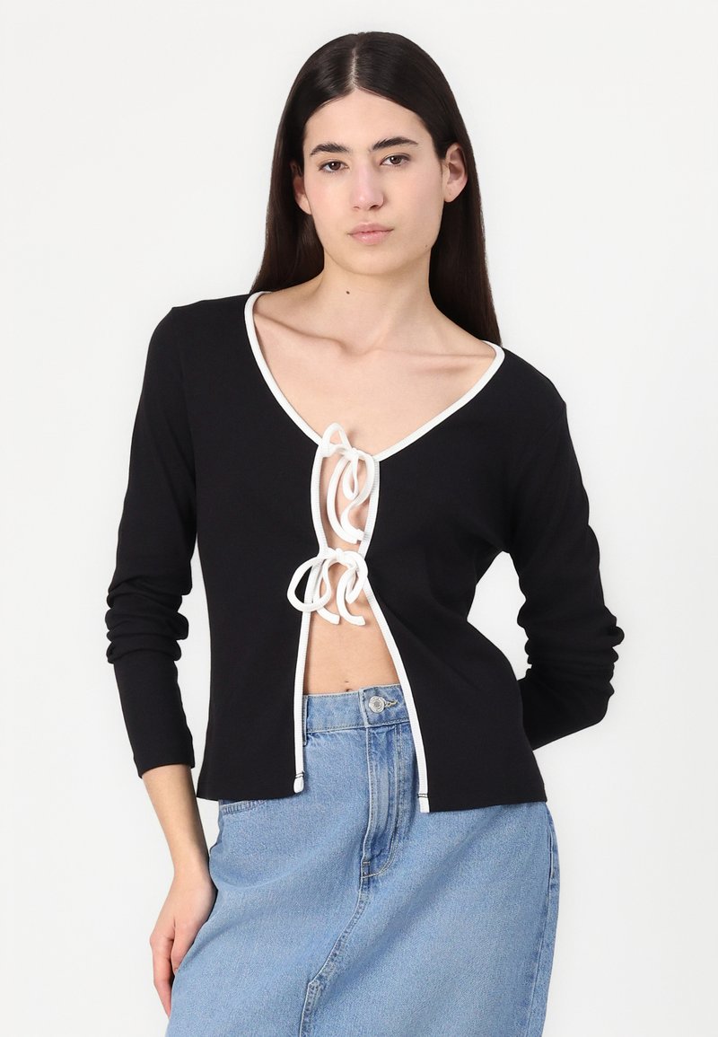 Top nero a maniche lunghe con scollo a V profondo, bordature bianche e chiusura a nodo frontale. Il tessuto sembra morbido; design complessivo aderente.