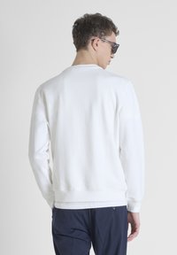 Felpa bianca in morbido tessuto, con collo rotondo classico e polsini a costine. La vista posteriore mostra un branding minimale e una vestibilità rilassata.