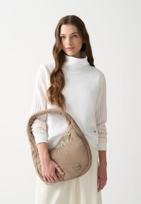 Barna, párnázott válltáska ívelt formával és széles pánttal, egy fehér, texturált ujjú felsőhöz tartva. Diszkrét logó részlet látható.