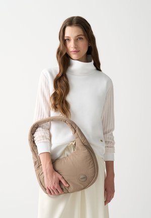 Brūna, mīksta plecu soma ar izliektu formu un platām siksnām, turot to pret balto topu ar teksturētām piedurknēm. Nedaudz pamanāma logotipa detaļa.