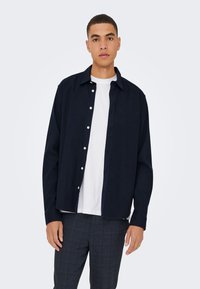 Temno modra srajca z dolgimi rokavi in belimi gumbi, nošena preko navadne bele majice, združena s sivimi karo hlačami.