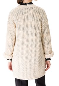 Personne portant un pull en grosse maille longue de couleur crème, avec les manches retroussées, par-dessus une chemise noire et blanche, vue de dos.