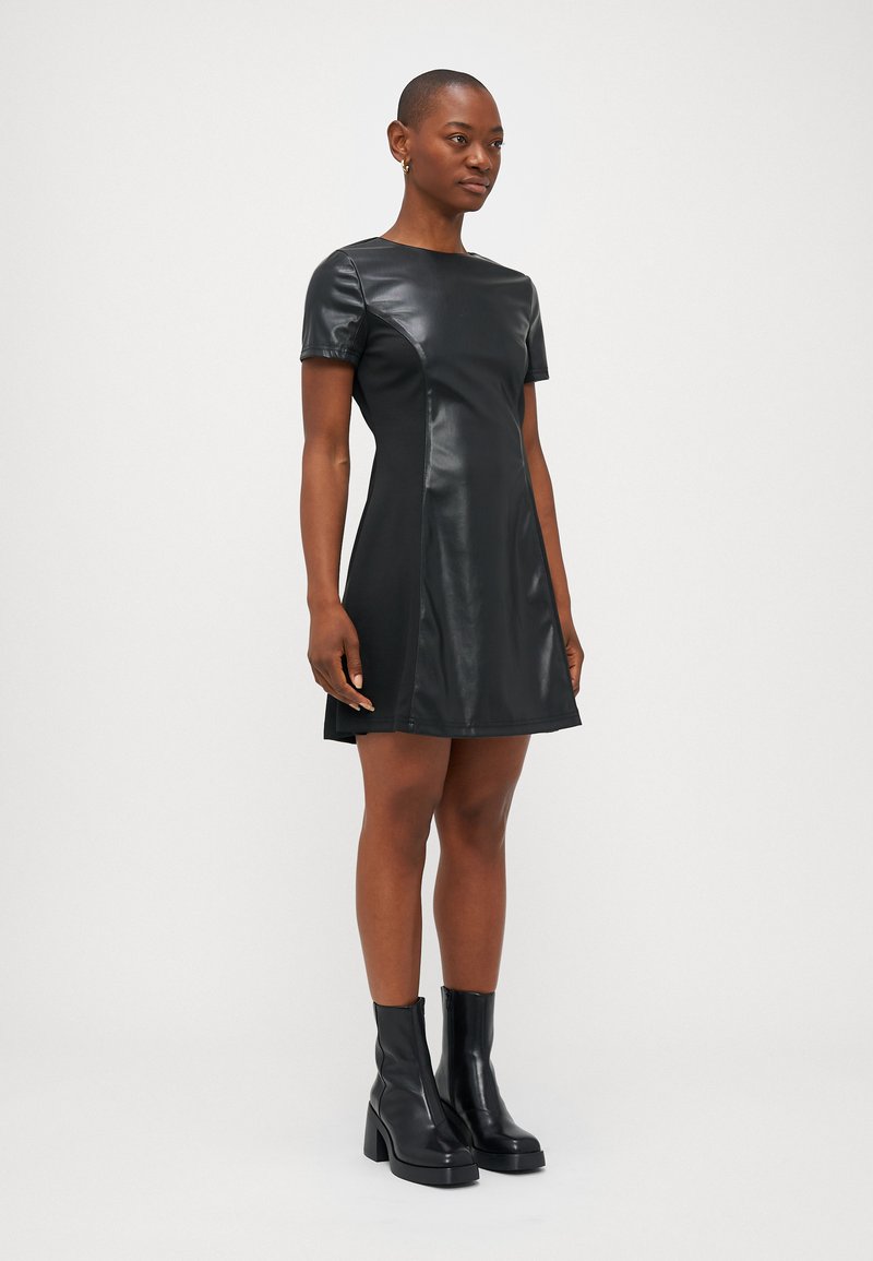 Schwarzes Kurzarmkleid aus Kunstleder mit tailliertem Oberteil und ausgestelltem Rock. Getragen mit schwarzen Absatz-Ankle Boots.