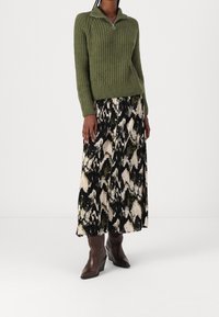 Suéter de punto acanalado verde con cuello con cremallera, combinado con una falda larga y estampada en negro, crema y verde, y botas de tacón marrones.