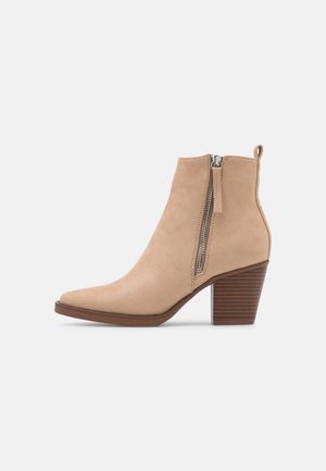 Bottine beige en daim avec bout pointu, fermeture éclair latérale, languette à l'arrière et talon bloc en bois empilé de hauteur moyenne.