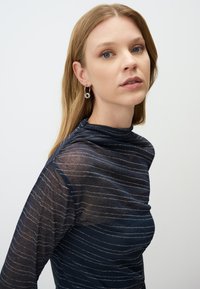 Top blu navy con un colletto alto, in tessuto trasparente e con strisce orizzontali argento. Modella che indossa orecchini a cerchio argentati, con il volto leggermente girato.