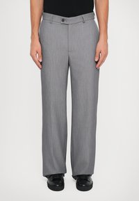 Man draagt rechte grijze pantalon met knoop- en ritssluiting, gecombineerd met zwarte sneakers en een zwart ingestopt shirt.