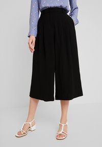 Kvinna i svarta vida cropped byxor, en blåmönstrad långärmad blus och vita sandaletter med blockklack.