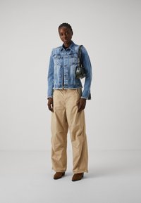 Denimjakke med knappdetalj og frynset kant, kombinert med beige, løstsittende bukser; fullfører antrekket med en liten mønstrete veske og brune støvler.