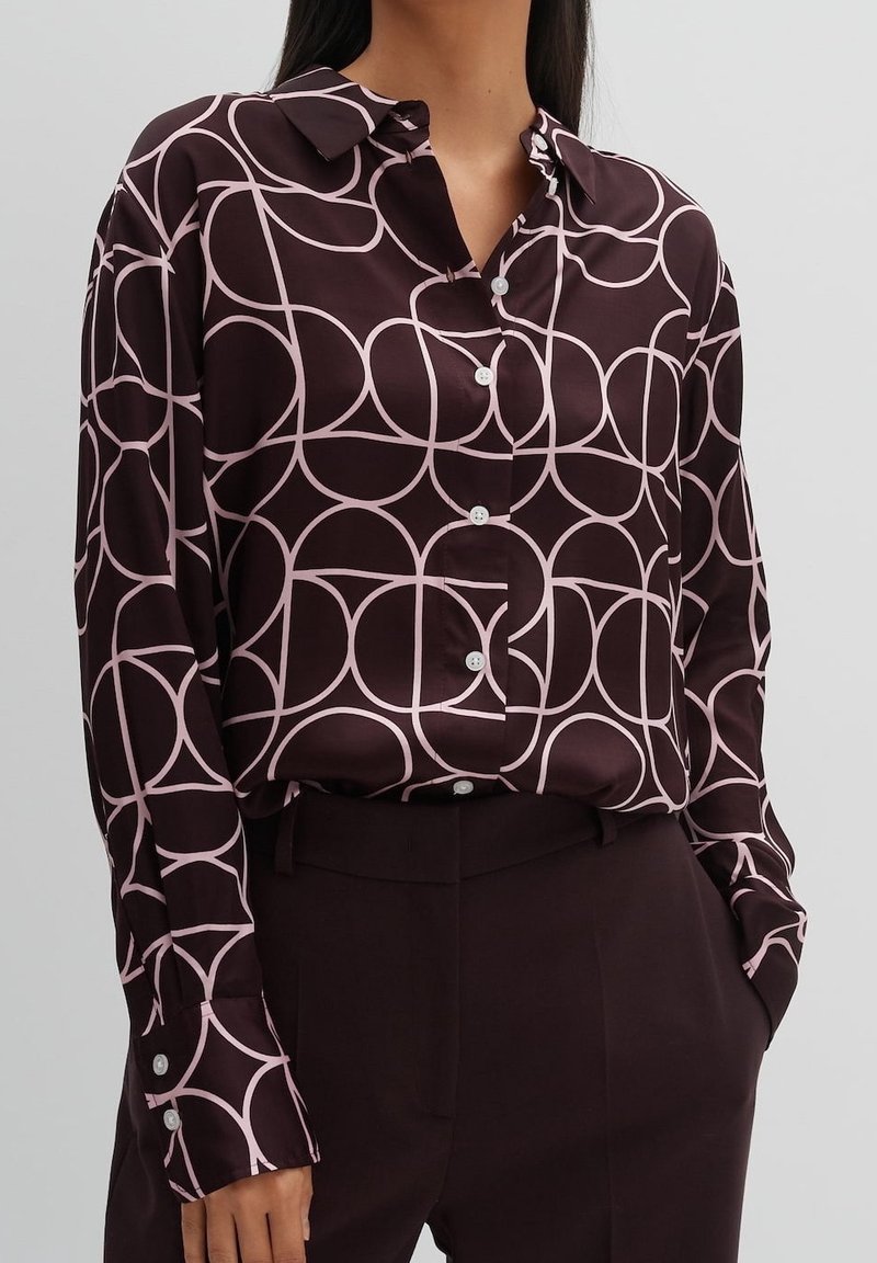 Femme portant une chemise boutonnée violet foncé avec un motif abstrait de cercles rose clair et un pantalon violet foncé, les mains dans les poches.