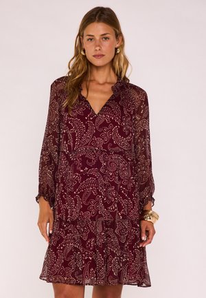 Donna che indossa un abito bordeaux con motivo floreale paisley, maniche lunghe trasparenti, scollo a V, orecchini a cerchio dorati e braccialetti dorati al polso destro.